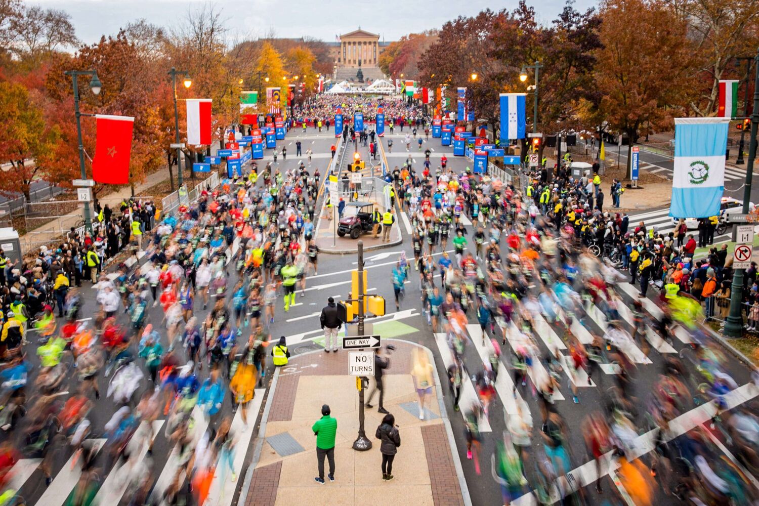 A long exposure shot of runners running, many being blurry due to motion. They're running down a street lined with different countries flags, with the Philadelphia Art Museum in the center background
